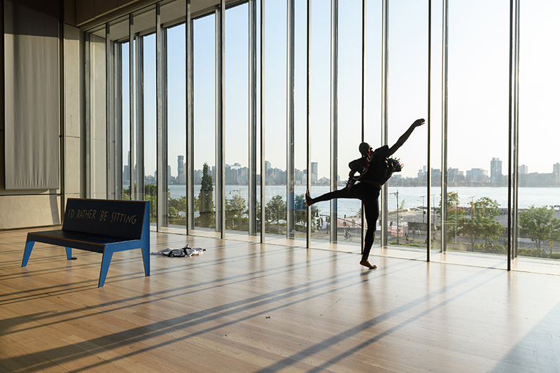 Color photo of Jerron in black leaping in the air against a wall of windows