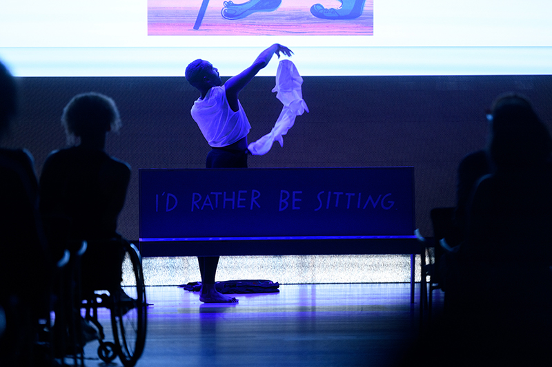 Color photo of Jerron shadowed in blue standing behind a bench undressing. A white shirt is suspended middair.