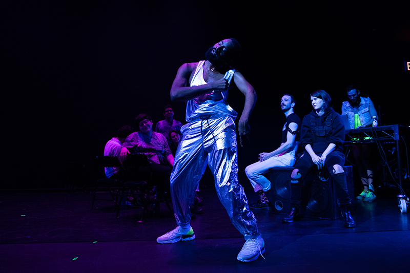 Color photo of Jerron dressed in a metallic suit swaying in front of audiences onstage