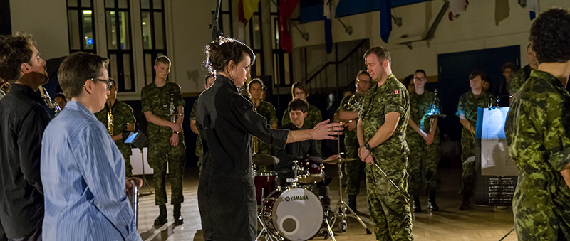 View of Marina Rosenfeld conducting the music performance in front of a group of military personnel in camo, a drummer, and the audience