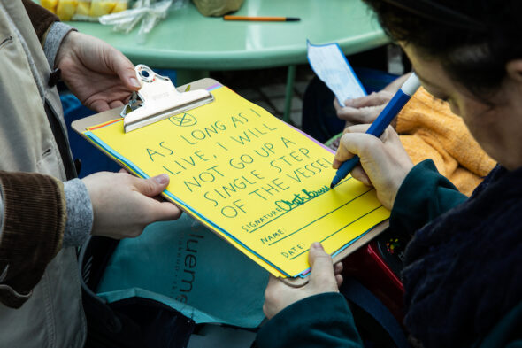 A close-up of Christine signing a pledge: “As long as I live, I will not go up a single step of the Vessel.” The pledge on colorful paper, riso-printed with blue hand-drawn text, and has a crossed-out-stairs symbol at the top.