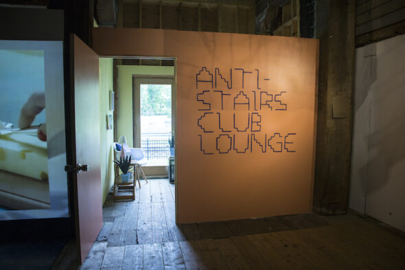 An orange wall with big text in a stair-inspired font that says, "Anti-Stairs Club Lounge."