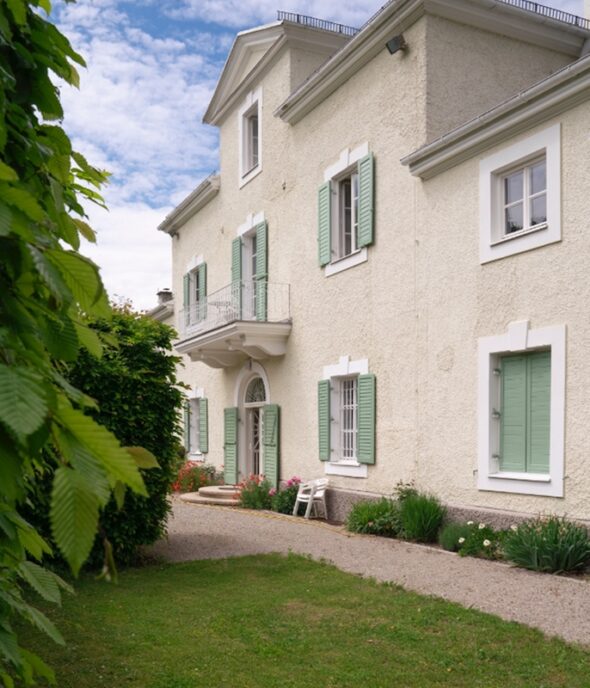 There is a white apartment building with two stories. The shutters are a seafoam green. There is a walkway in front of the building as well as grass and a small tree.