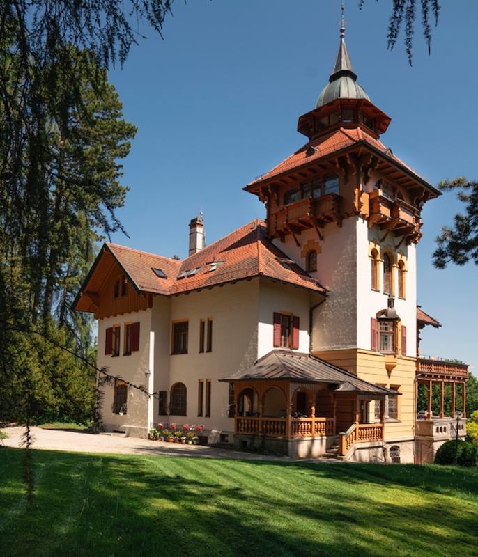 A traditional Alpine house with wooden shutters, a wood terrace and a spiraling turret is situated on a plot of lush green grass. Leaves from surrounding trees and a cloudless blue sky frame the image.