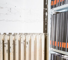 an abstract, close up photograph of an old and slightly damaged white radiator next to a shelf with a dozen black archival binders, each with an orange label on it