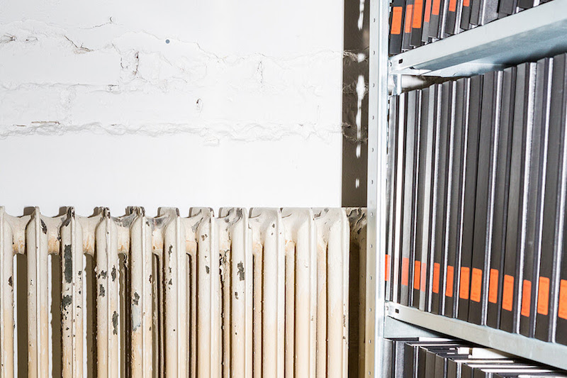 an abstract, close up photograph of an old and slightly damaged white radiator next to a shelf with a dozen black archival binders, each with an orange label on it