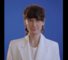 a still from a video portrait of the artist Manuel Solano wearing a white suit jacket over a white shirt, against a blue background, gaze focused on the viewer
