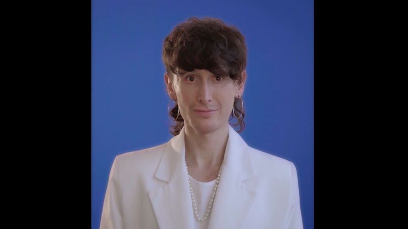 a still from a video portrait of the artist Manuel Solano wearing a white suit jacket over a white shirt, against a blue background, gaze focused on the viewer