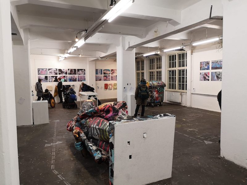 a large gallery space with bright white lights, an installation with coloured textiles in the foreground and photos mounted on the walls, several people looking at them, some standing, some in wheelchairs