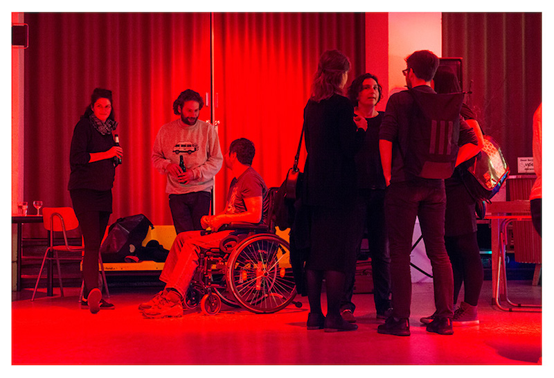 in a red-lit room with a curtain against the back wall, a group of people stand or sit in wheelchairs, speaking to each other and having a drink