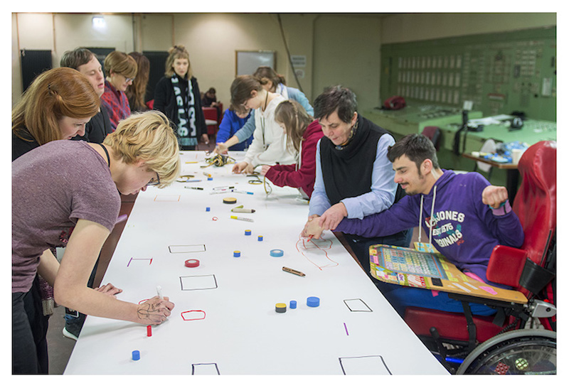 a group of artists stands or sits in wheelchairs around a long table with a white paper on it, drawing different and moving small pieces around