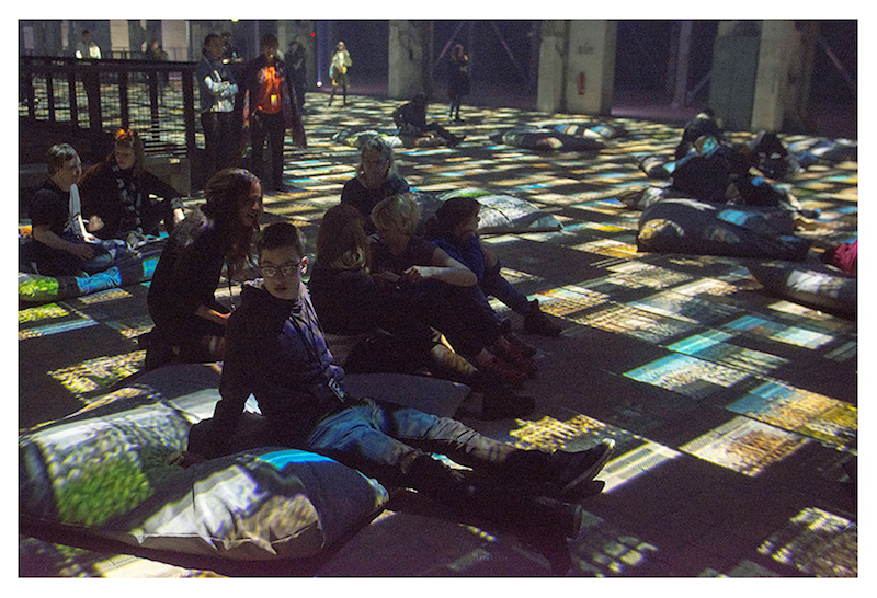 a group of people sit on the floor on pillows, as a display of colorful lights surrounds and covers them