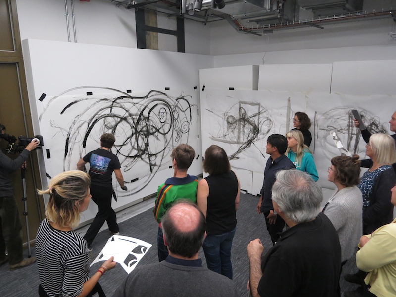 participants in a workshop for partially sighted and blind people in architecture pictured in front of two large wall canvases, with the teacher drawing black circular figures on them