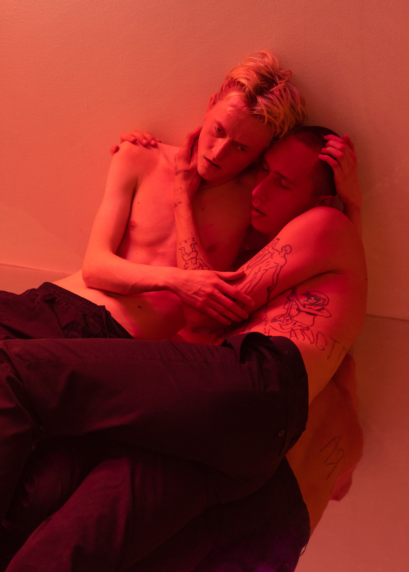 two shirtless people are hugging, leaning against a wall, bathed in red light