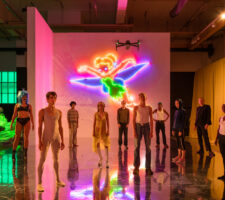 ten young performers are standing on a stage with mirrored floor and pink lighting, the light comes mostly from a large neon Tinkerbell mounted on a white wall behind them