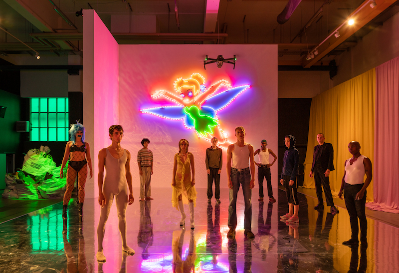 ten young performers are standing on a stage with mirrored floor and pink lighting, the light comes mostly from a large neon Tinkerbell mounted on a white wall behind them