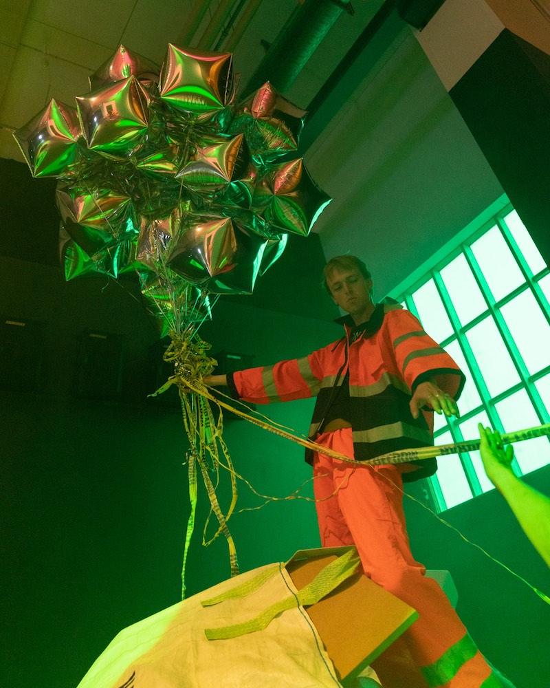 A person in hi-vis construction suit leans against a gridded window, while holding a large bunch of silver helium balloons