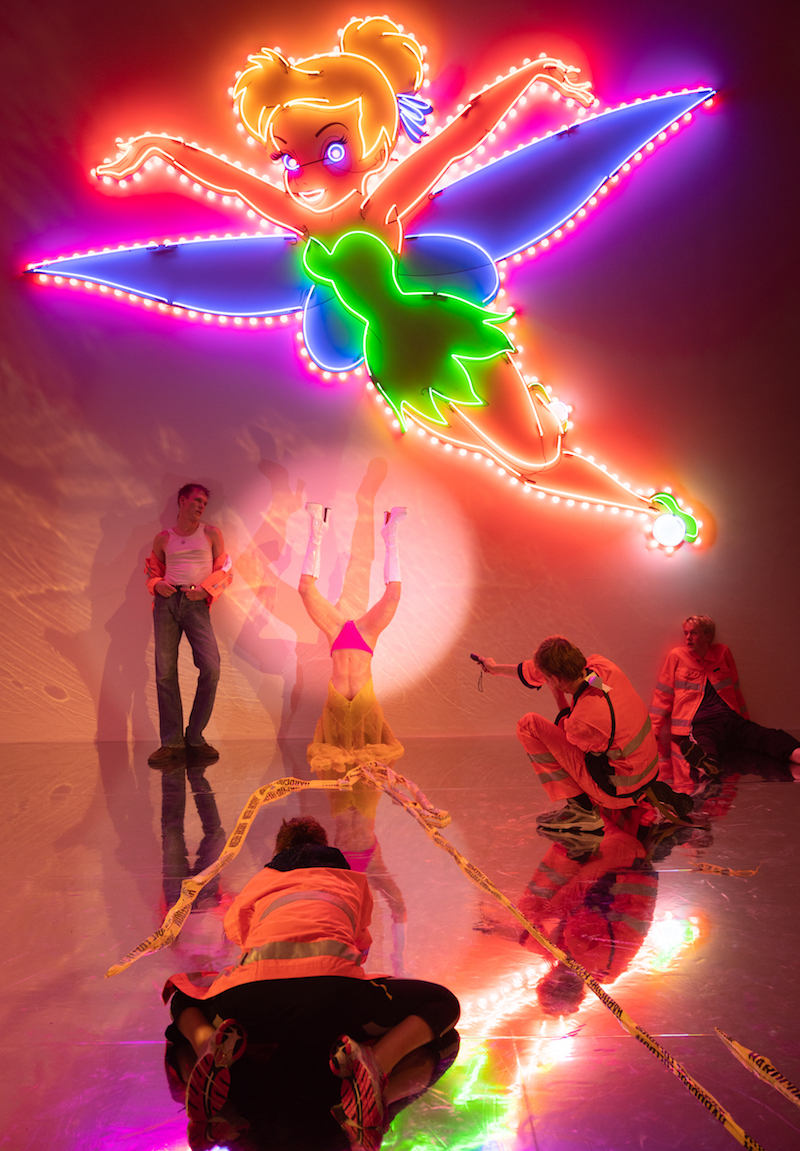 under a neon mounted tinkerbell light, several performers are dancing or draped on a mirrored stage