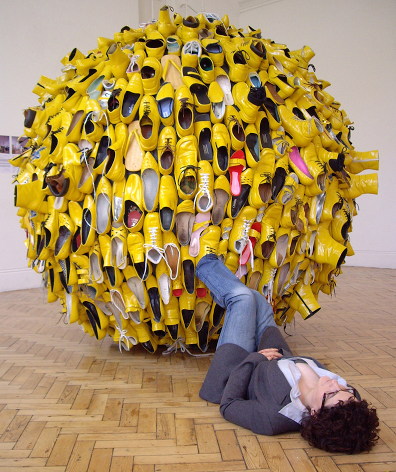 a person lies down on the floor of a gallery with her feet in the air, one foot is placed inside a shoe that forms part of a large, yellow, globe sculpture made up of shoes