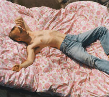 a shirtless person with a shaved head and wearing only pale blue jeans lies on a bed, on top of a rose coloured, flower pattern duvet