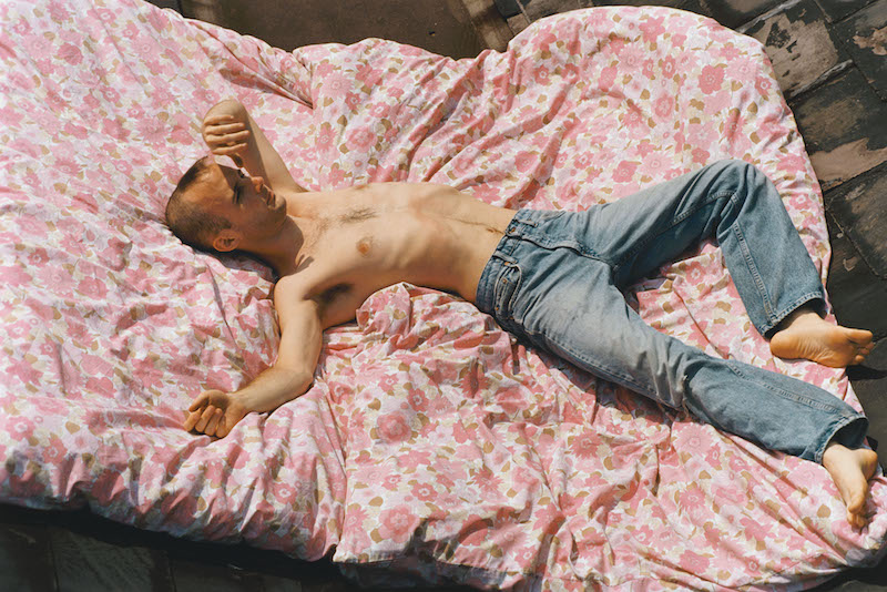 a shirtless person with a shaved head and wearing only pale blue jeans lies on a bed, on top of a rose coloured, flower pattern duvet