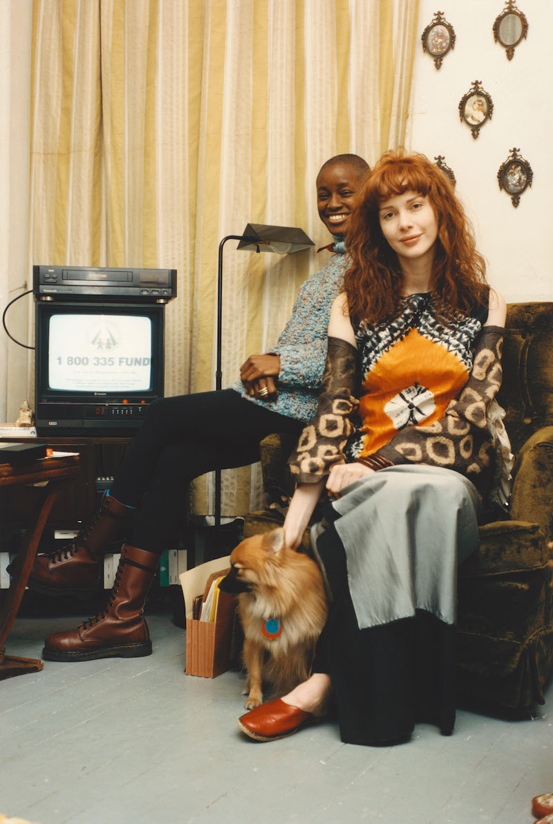 two people sit on a lazy boy next to an old television set, with a small dog between them on the floor