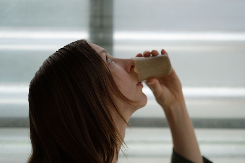 A woman tips her head backwards to drink from a small cup made from algae-based bioplastic. She holds the cup to her lips with her left hand.