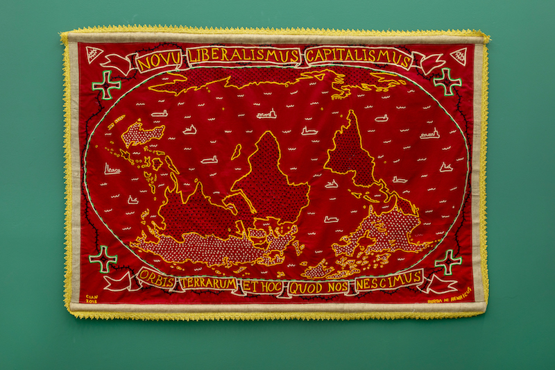 A detailed shot of a tapestry, embroidered with a map reading "novu imperialismus capitalismus" across the top is mounted on a green gallery walll