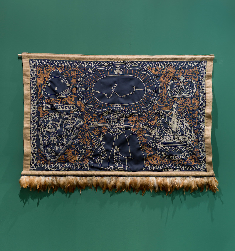 an embroidered textile artwork in deep navy and rust colour, showing an unknown map, is mounted on a forest green gallery wall