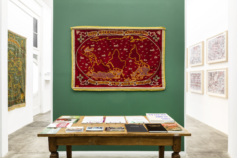 a red textile embroidered map, hard to make out in detail, is mounted on a forest green gallery wall. a table of literature sits in front of the artwork