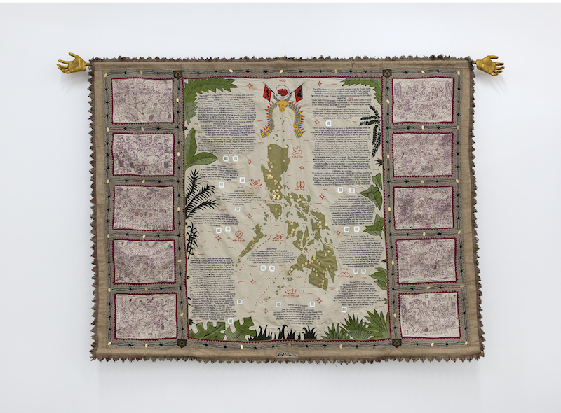 an embroidered textile map with writing on it is mounted on a white gallery wall