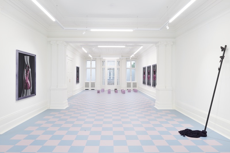 a wide angle shot of a gallery space with works mounted on the wall and a pale blue and pink checkered floor