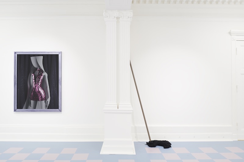 a portrait mounted on a white gallery wall, beside an oversized black mop, which leans against the wall