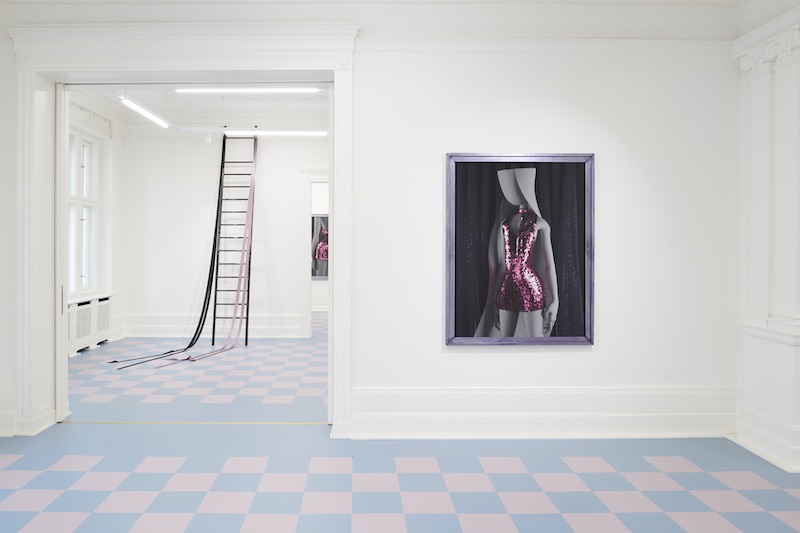 a portrait of a girl in a sequined dress mounted on a white gallery wall, with a ladder coming down from the ceiling in the background