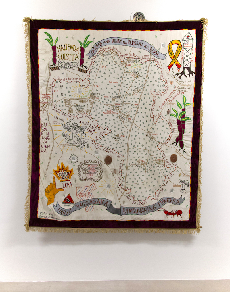 a colourful embroidered tapestry showing an unknown map is mounted on a white gallery wall