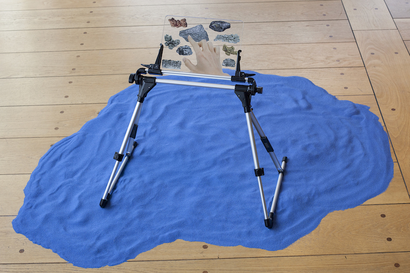 a small stand holding a transparent plaque with images of different minerals on it, sits on top of a pile of bright blue sand