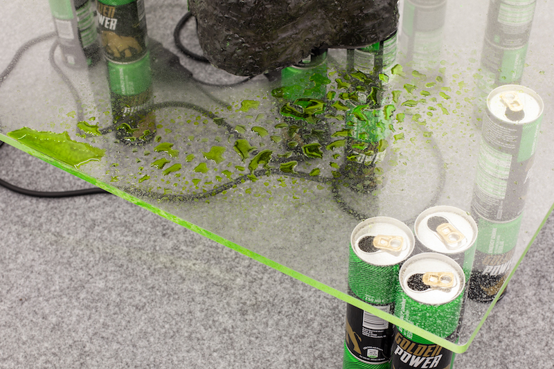 a transparent table is partially shown, held up with cans of green energy drinks instead of table legs, a mass of cords under it. there is a spilled green liquid on the table top