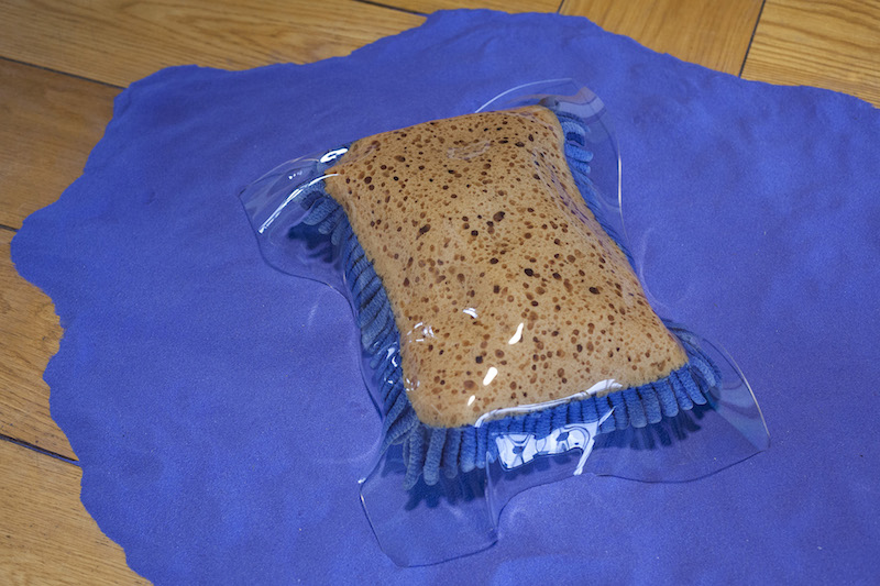 a pile of bright blue sand on a floor, with a large sponge sitting atop it, encased in clear plastic