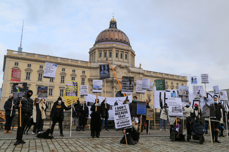 protest: group of cultural workers in smocks with posters reading 'i don't participate because' standing in front of Humboldt forum berlin