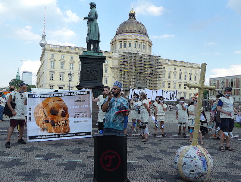 A man gives a speech in front of a group of protestors, standing next to a scultpture of a cross and orb, like the one on top of humboldt forum