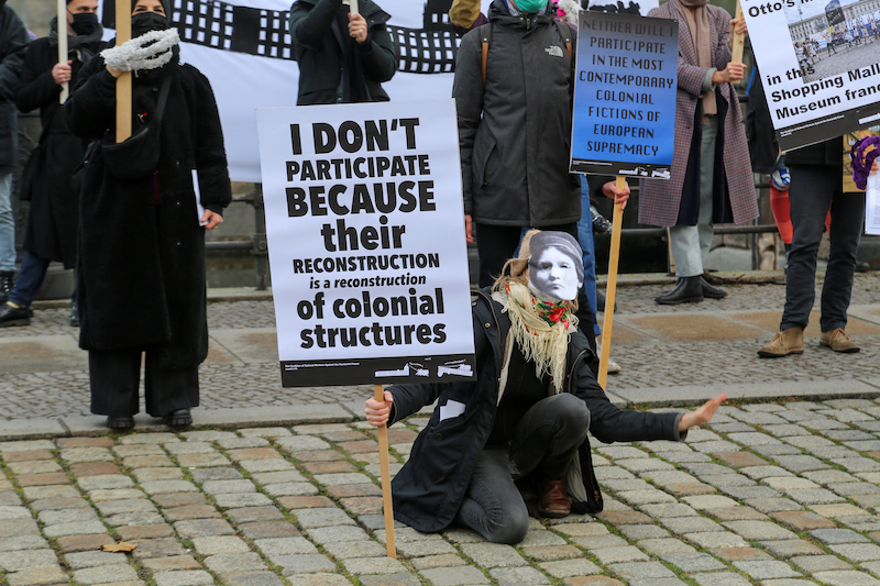 A person wearing a face mask kneels while holding picket sign 'I don't participate because their deconstruction is a reconstruction of colonial structures'