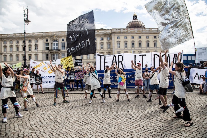 a group of protestors in smocks hold flags and signs in a half circle and have their arms up, performing a song