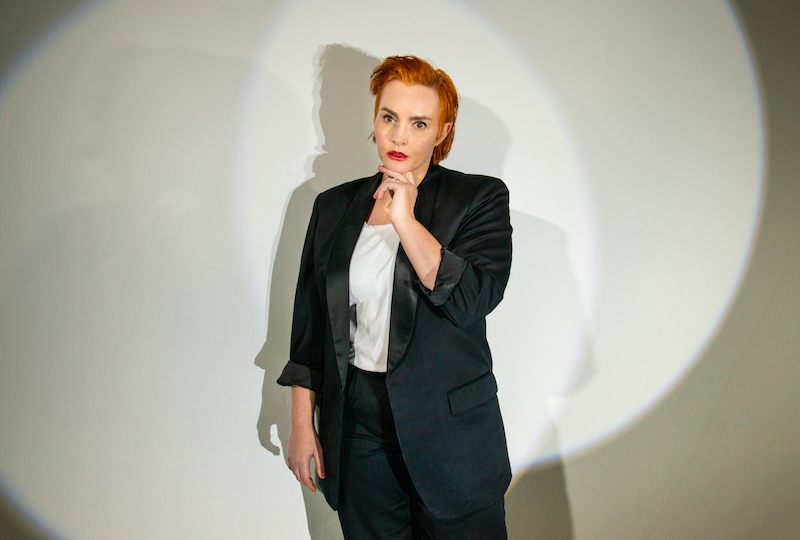 a person with short, sleek orange-colored hair stands in front of a circular stage light, wearing a black lounge suit and white shirt, she looks into the camera