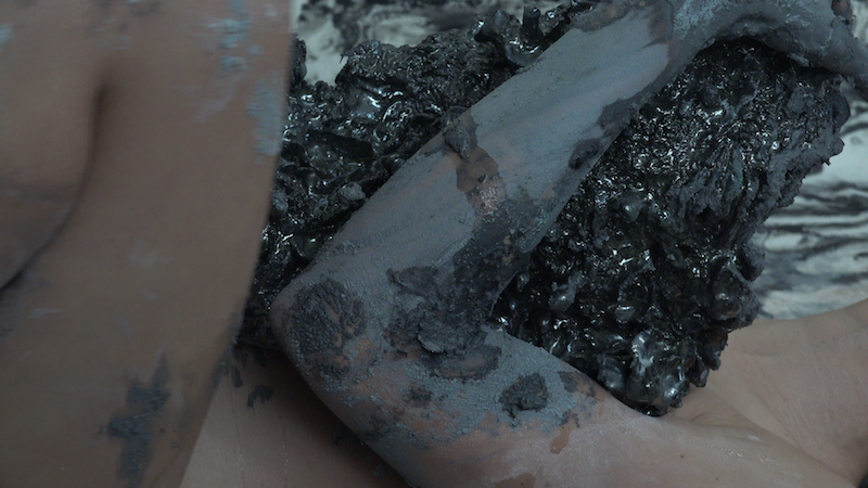 a close up still from a video, featuring some unidentifiable limbs covered in dark clay