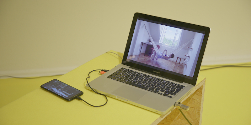  laptop and phone are sitting on a yellow bench depicting the artist in a hammock.