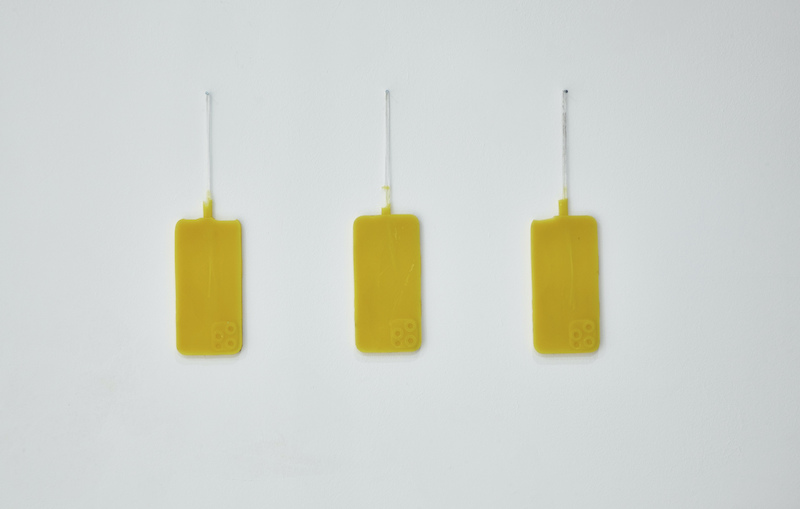 Three yellow beeswax pressings of iPhones hanging on a white wall.