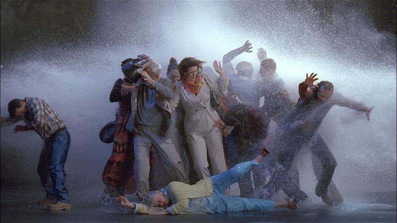 a video still of a group of people in a storm, a tableau that looks as if the people are being pushed and toppled by the wind
