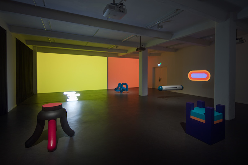 an installation view of a gallery space with yellow and orange lit walls, showing several abstract geometric sculptures across the floor