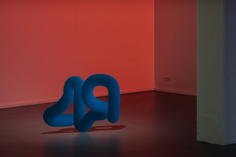 a squiggly blue sculpture sits on the floor in front of a red-list wall