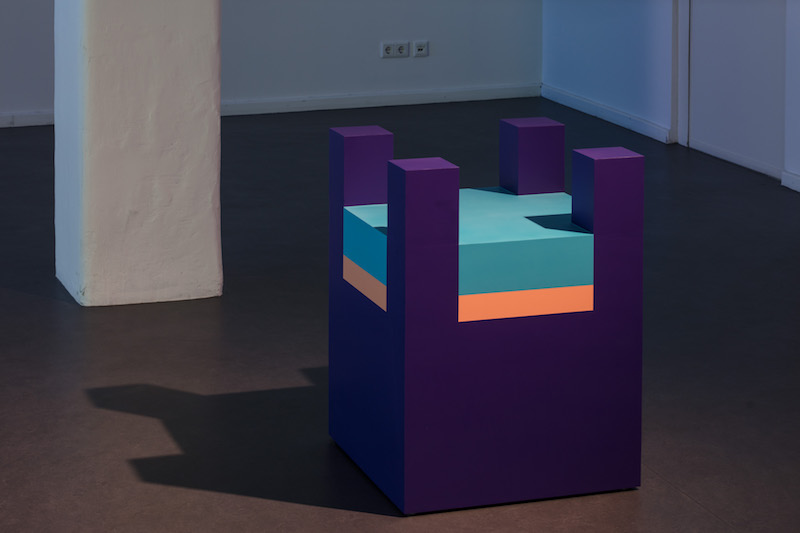 a boxy purple, pink and blue sculpture resembling a lego block sits on a concrete gallery floor